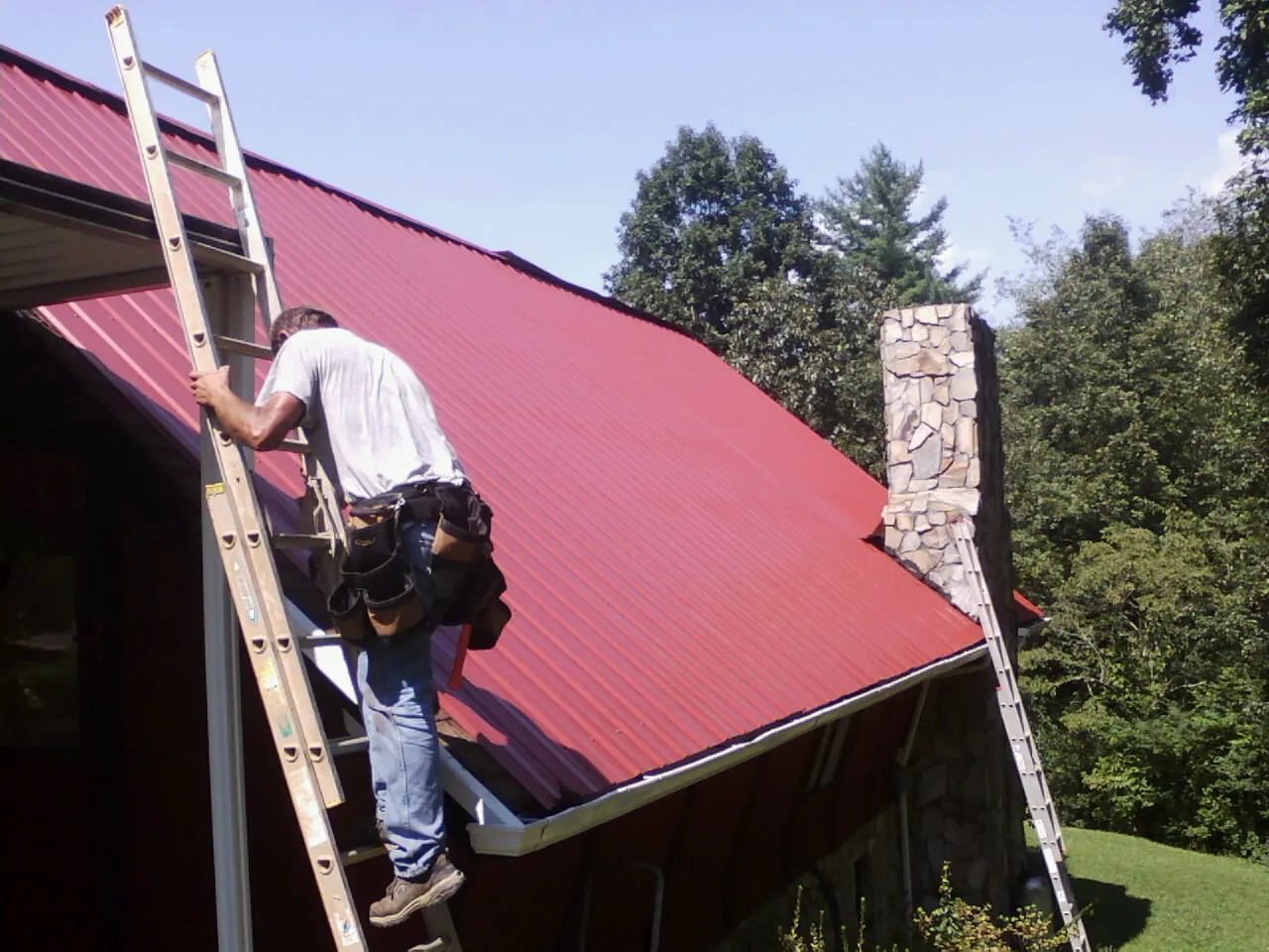 Roofer on a ladder beside a red metal roof and stone chimney — Soffit & Fascia Repair by Roofing Brothers in Paynesville